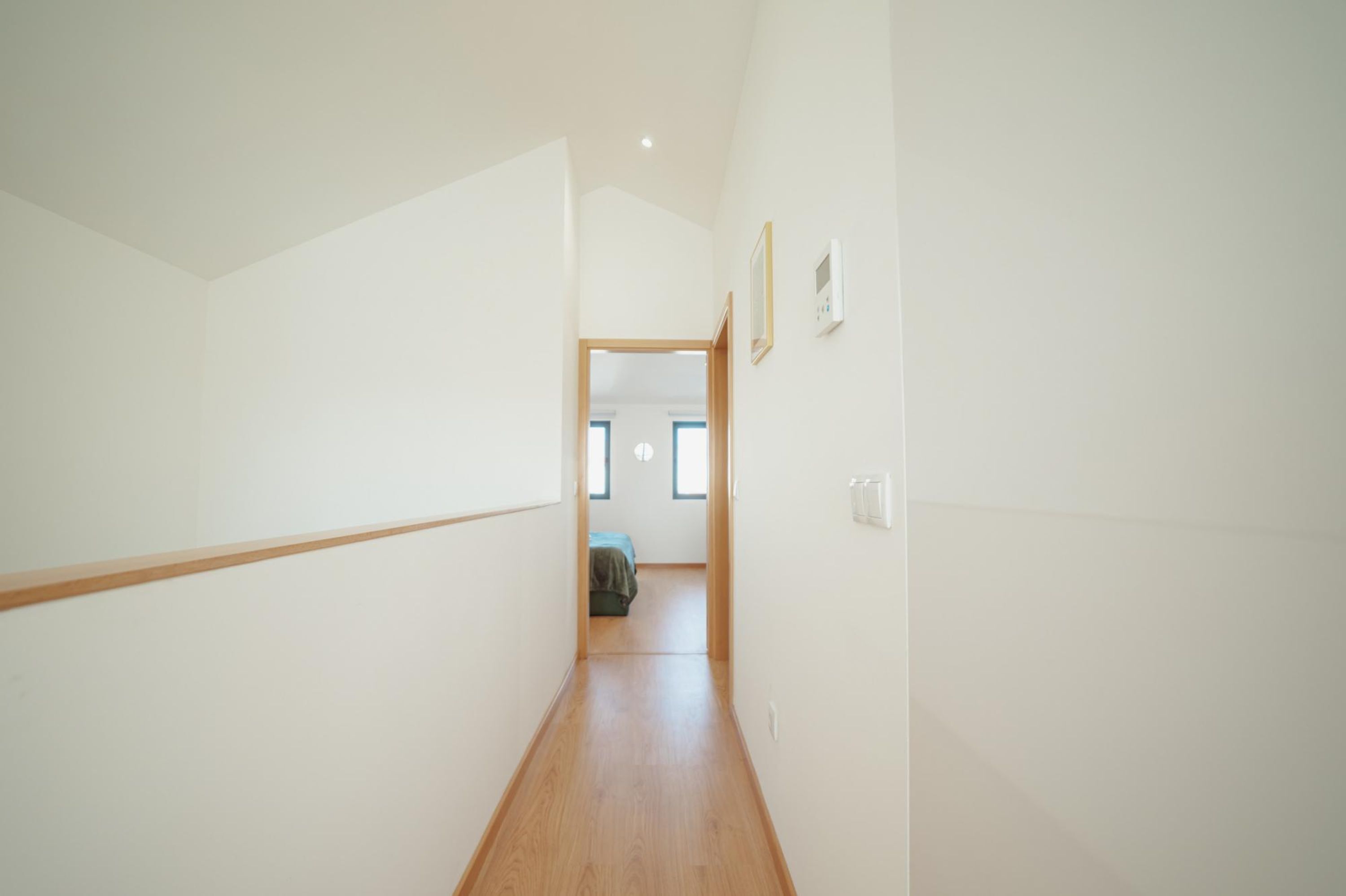 Un couloir étroit et bien éclairé, doté d’un plancher en bois et de murs blancs, qui mène à une chambre à coucher avec un lit et une fenêtre.