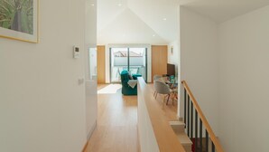 A modern interior with a wooden staircase, a dining area with a table and chairs, and a living space with a sofa and a window.