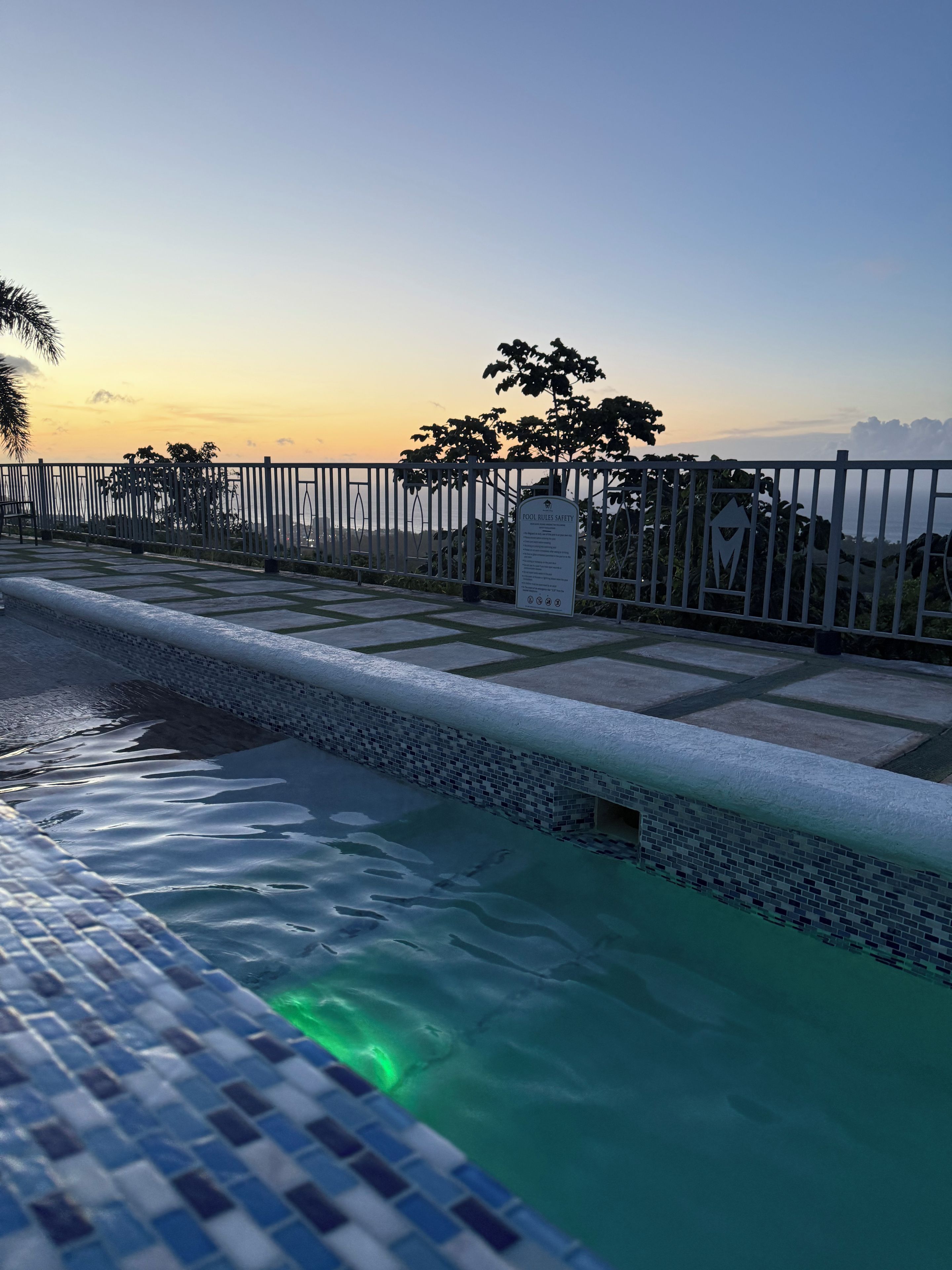 Una piscina con borde de azulejos, un letrero de seguridad y una valla con árboles al fondo.
