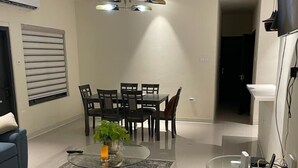 Ein modernes Wohnzimmer mit Essbereich, ausgestattet mit einem blauen Sofa, einem Glastisch mit Pflanze, einem Esstisch mit Stühlen und einer außergewöhnlichen Deckenleuchte.