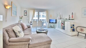 A modern living room with a beige sofa, a glass coffee table, and a flat-screen TV. There is a balcony with a view of the city. - Seebrücke Appartement 14 - Seebrücke (Scharbeutz)