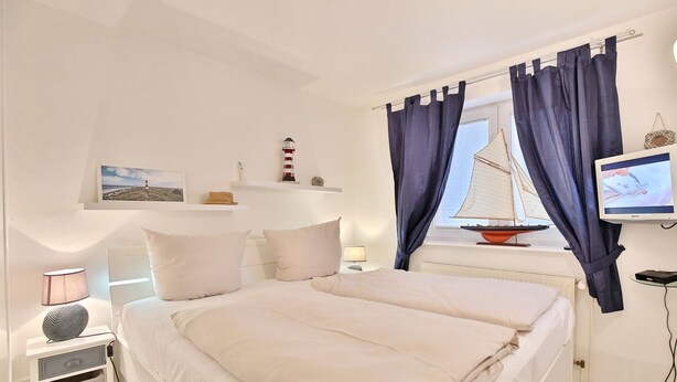 A bedroom with a bed, a nightstand, a lamp, a television, and a window with curtains. - Seebrücke Appartement 14 - Seebrücke (Scharbeutz)