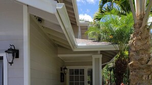 Uma casa com fachada branca, varanda de madeira, uma palmeira e um pequeno jardim com grama sintética e pedras.