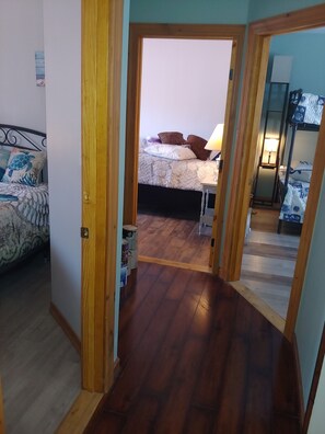 A hallway leading to a bedroom with a bed, a nightstand, and a lamp. There is a wooden floor and a door on the left. - Black Iron beach house (Blossvale)