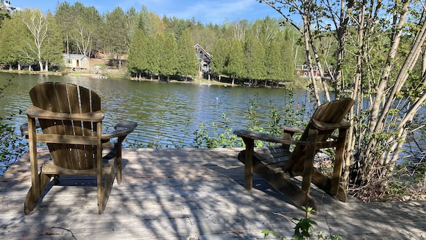 Una terrassa de fusta amb dues cadiretes Adirondack orientades cap a un llac tranquil, envoltades d'arbres i una petita cabana al fons.