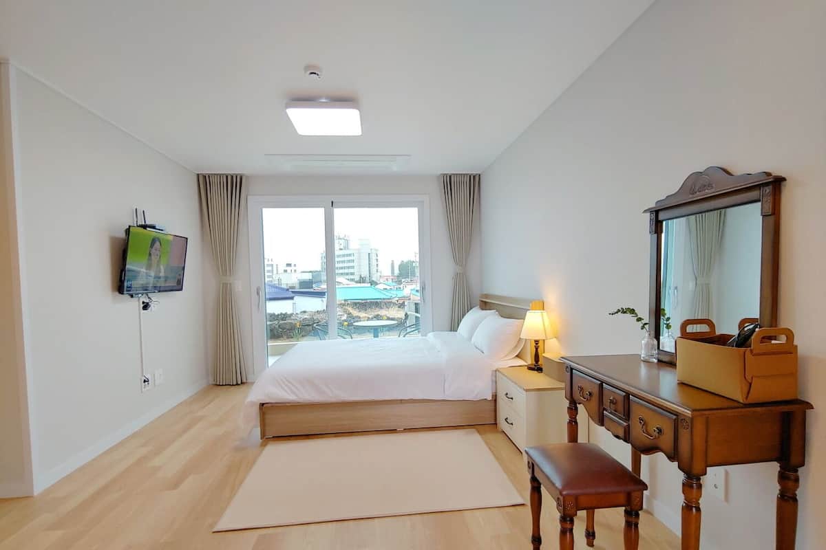 Habitación de hotel con cama, mesita de noche, lámpara, espejo, banco de madera y televisor montado en la pared.