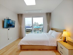 A hotel room with a large bed, a TV mounted on the wall, a bedside table with a lamp, and a view of the city through the window. - Viravi Stay (Jeju City)