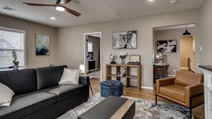A modern living room with a black sofa, a blue ottoman, and a wooden coffee table. There is a ceiling fan, a TV, and a bookshelf with decorative items. - The Altitude Loft (Bartlesville)