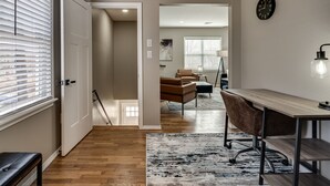 A modern living room with a wooden floor, a large window with blinds, a black pendant light, a desk with a chair, and a clock on the wall. - The Altitude Loft (Bartlesville)