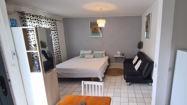 Un dormitorio pequeño con cama, sofá negro, mesita y refrigerador blanco.