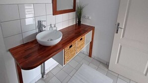 Un baño con tocador de madera, azulejos blancos, un lavabo blanco, un espejo y una puerta.