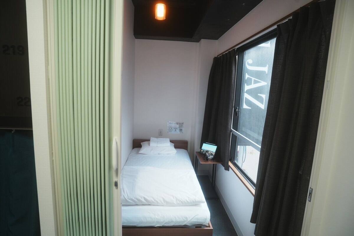 A small, clean room with a bed, a window with curtains, and a small shelf with a camera.