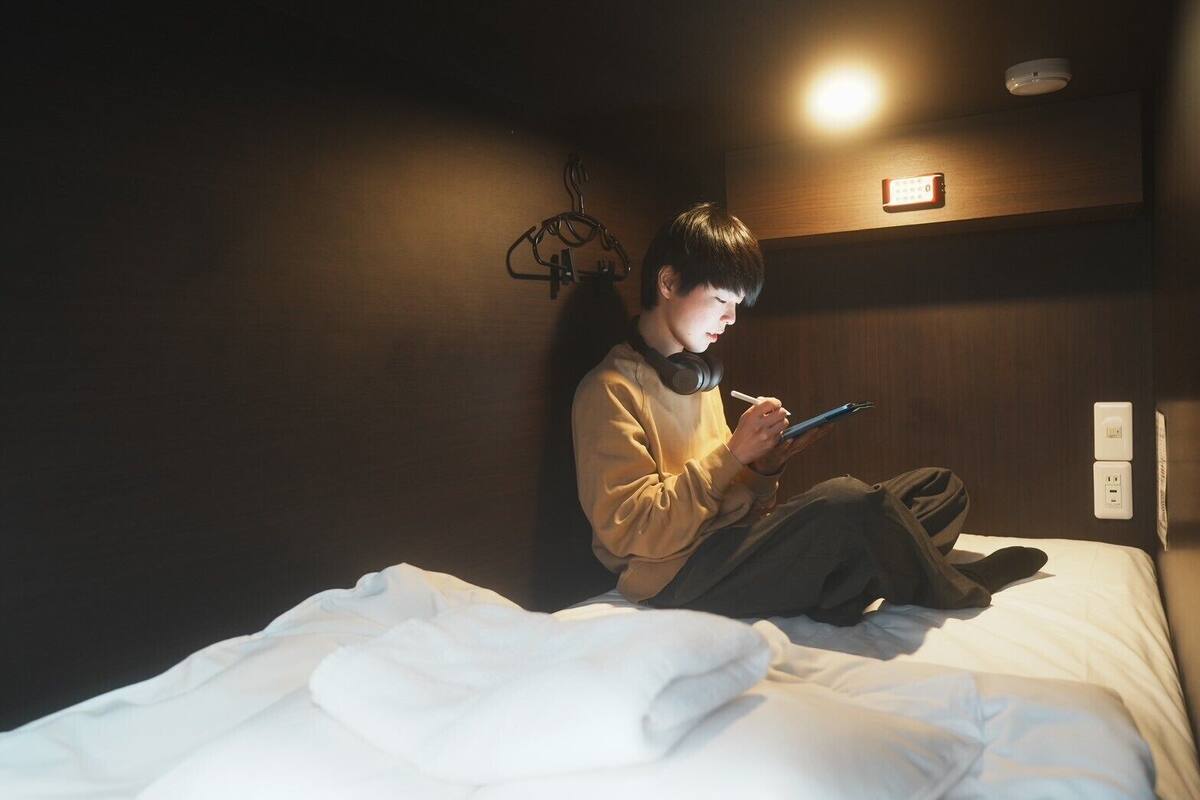 A person sitting on a bed in a dimly lit room with a wall-mounted light, a power outlet, and a small shelf with a remote control.