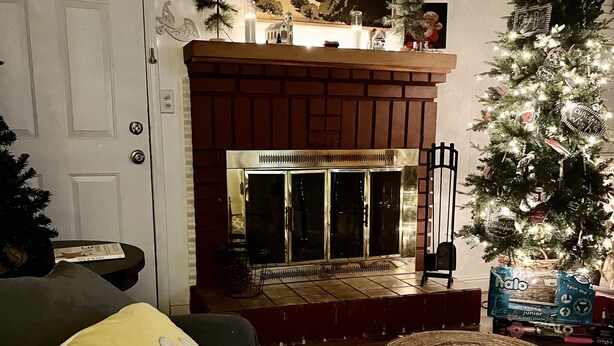 Un salon avec une cheminée, un sapin de Noël et un canapé agrémenté d’un coussin orné de la mention « Joyeux Noël ».