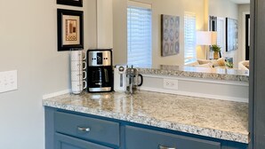 A kitchen with a granite countertop, a coffee maker, a kettle, and a coffee pot. There are framed pictures on the wall and a window with blinds. - Grand Rapids Dayton Station! (Grand Rapids)