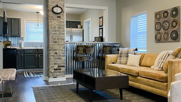 A modern living room with a sofa, coffee table, and a rug. There is a kitchen in the background with a refrigerator and cabinets. - Grand Rapids Dayton Station! (Grand Rapids)
