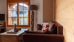 Ein gemütliches Wohnzimmer mit Holzwand, einem großen Fenster mit Blick auf Gebäude, einem braunen Sofa, einem Holztisch mit Früchten und einer Lampe.