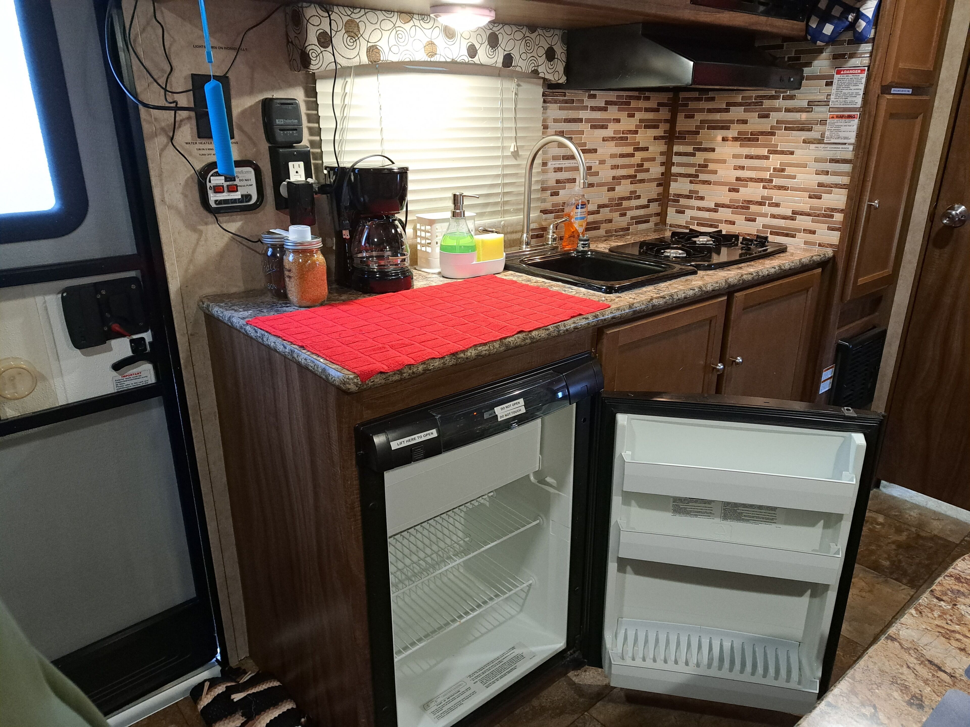 A compact kitchen with a dishwasher, a sink, and a stove. There is a red mat on the countertop, and various kitchen items are visible.
