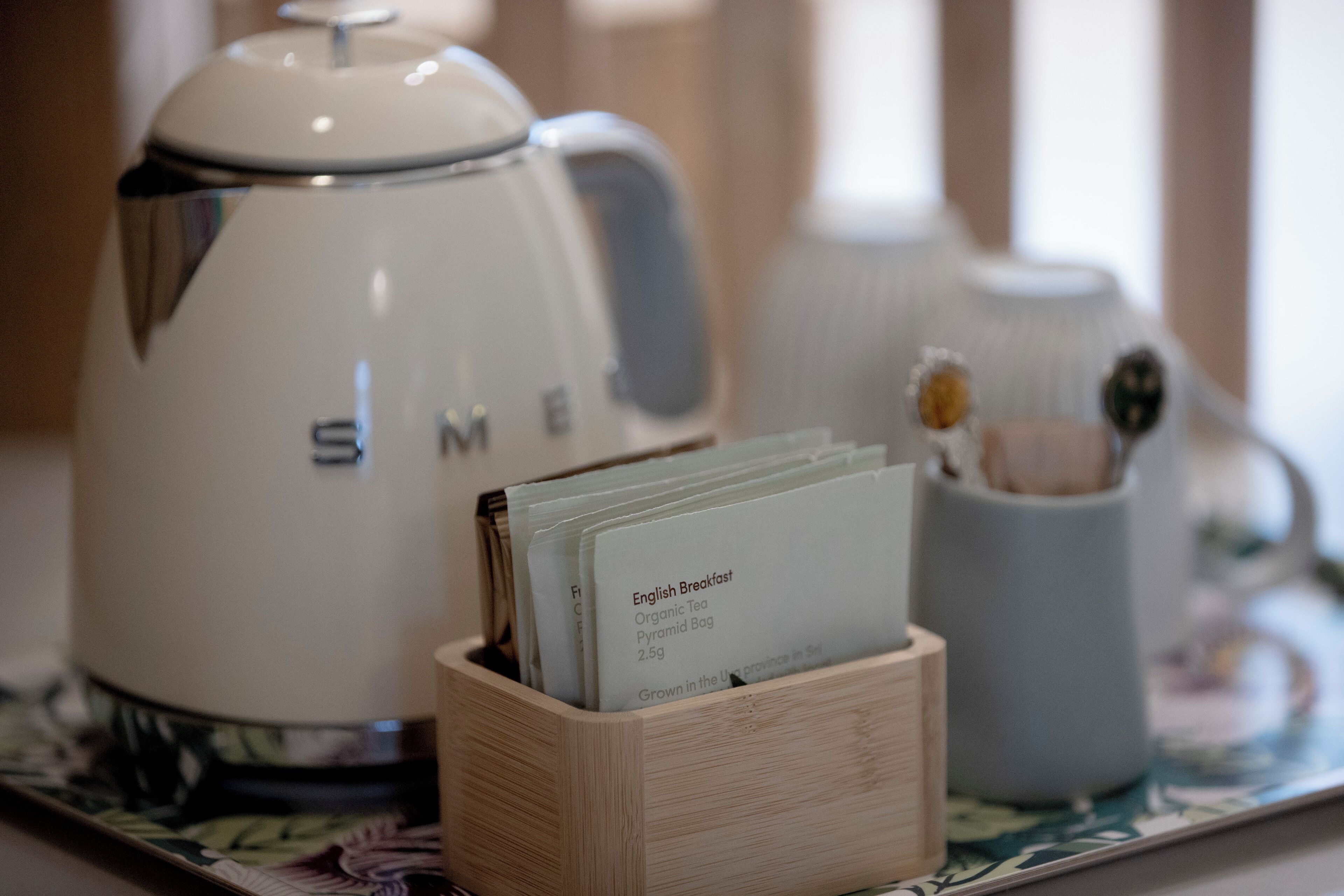 A white kettle with the brand name "SME" on a tray with a wooden block holding a tea bag labeled "English Breakfast Organic Tea."