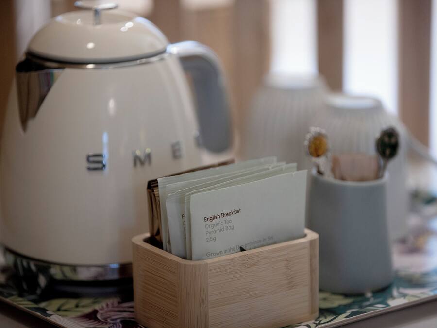 Una tetera blanca con el nombre de marca "SME" sobre una bandeja con un bloque de madera que sostiene una bolsita de té etiquetada como "Té Desayuno Inglés Orgánico".