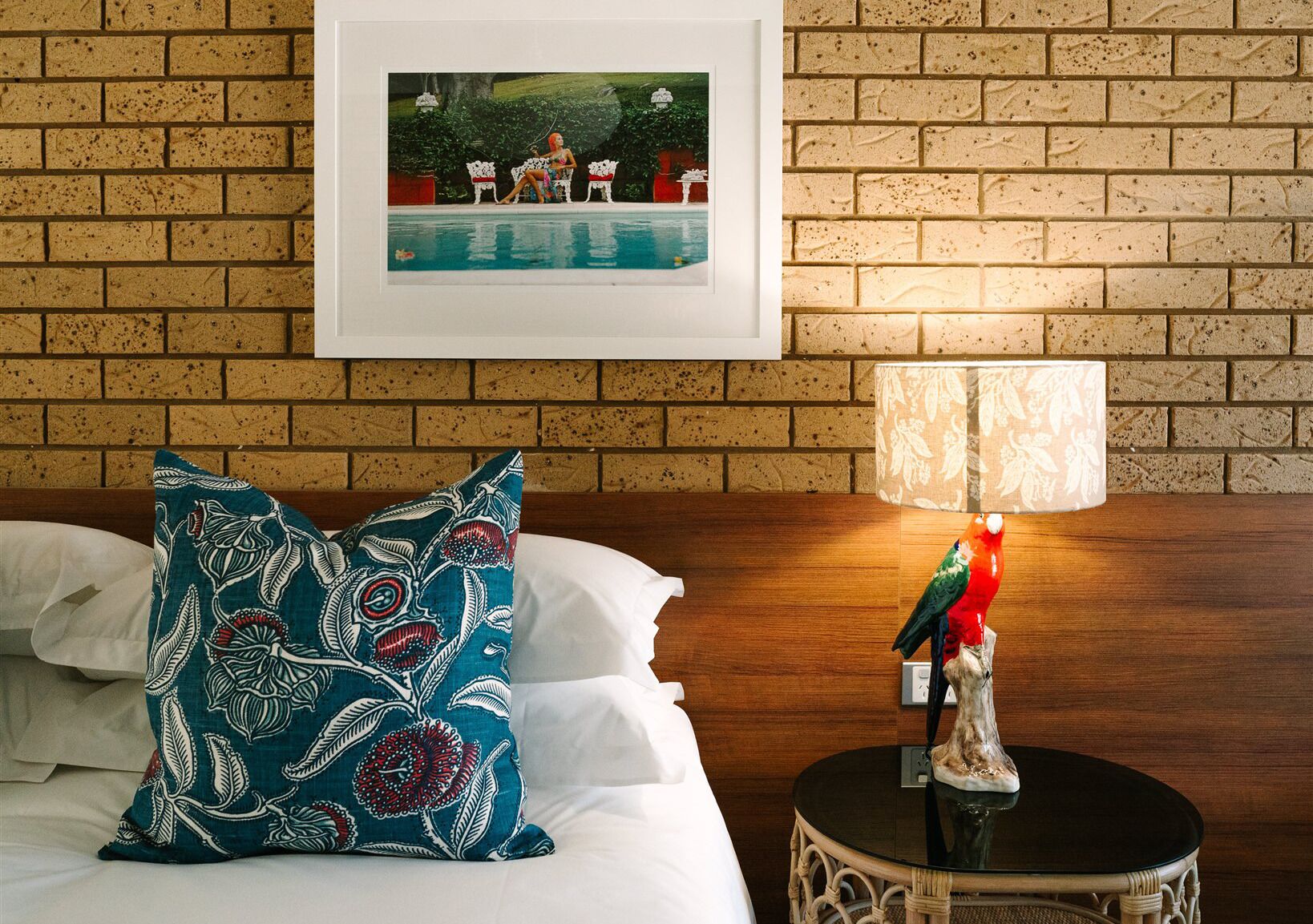 A hotel room with a brick wall, a framed picture of a pool, a white bed with a teal patterned pillow, and a table lamp with a bird figurine.
