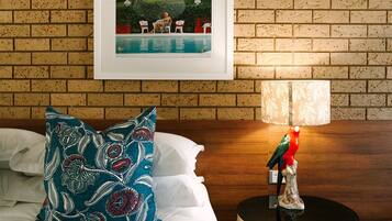A hotel room with a brick wall, a framed picture of a pool, a white bed with a teal patterned pillow, and a table lamp with a bird figurine.