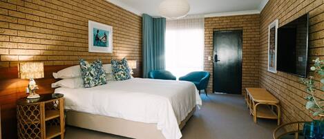 A hotel room with a brick wall, a bed with white linens, a blue chair, a small table, and a TV mounted on the wall.