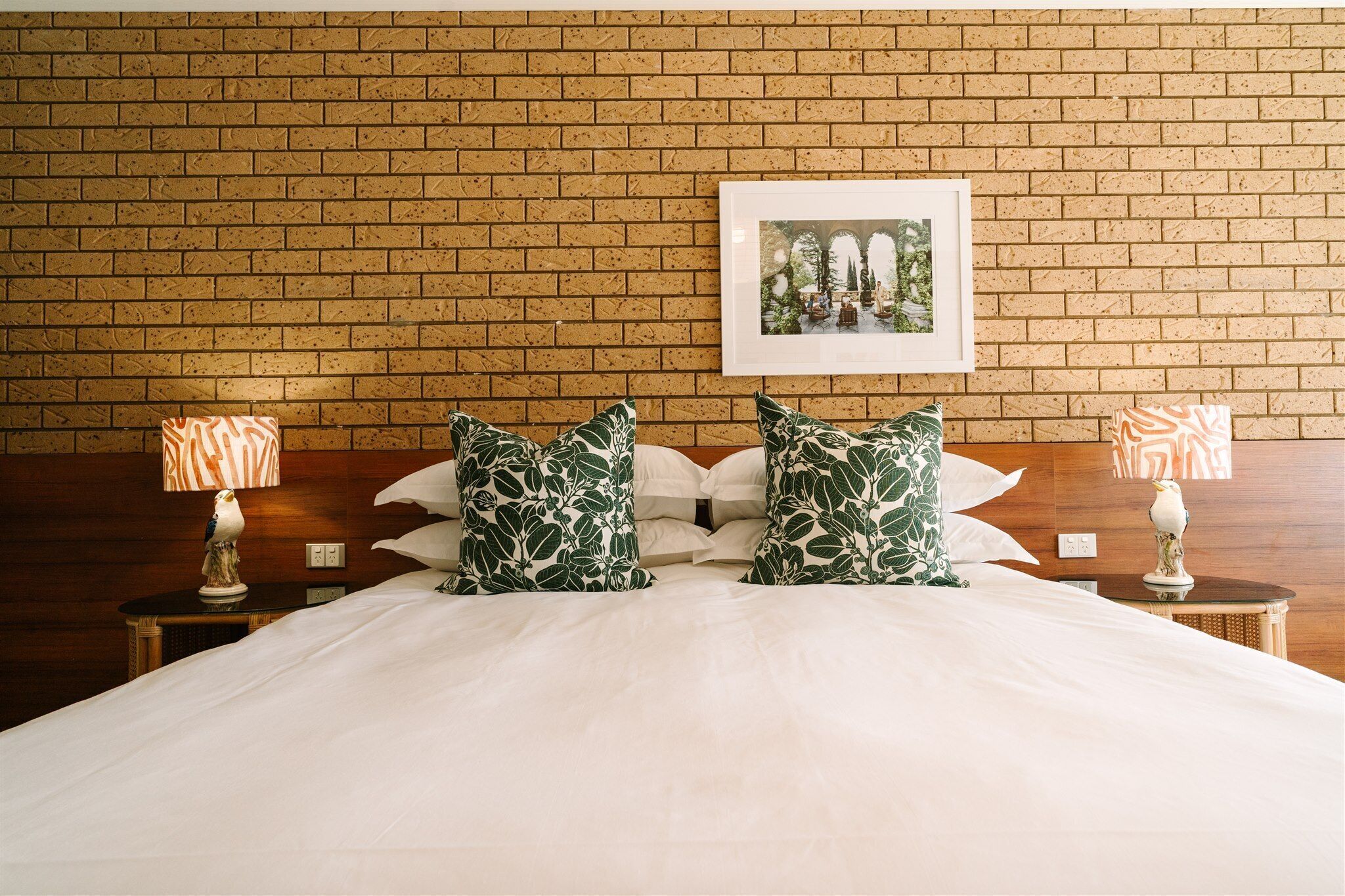 Una habitación de hotel con pared de ladrillo, un cuadro enmarcado, una cama con ropa blanca y dos mesitas de noche con lámparas.