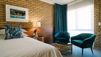 Een hotelkamer met een bakstenen muur, een bed met opvallende kussens, een nachtkastje met lamp, een stoel en een groot raam met gordijnen.
