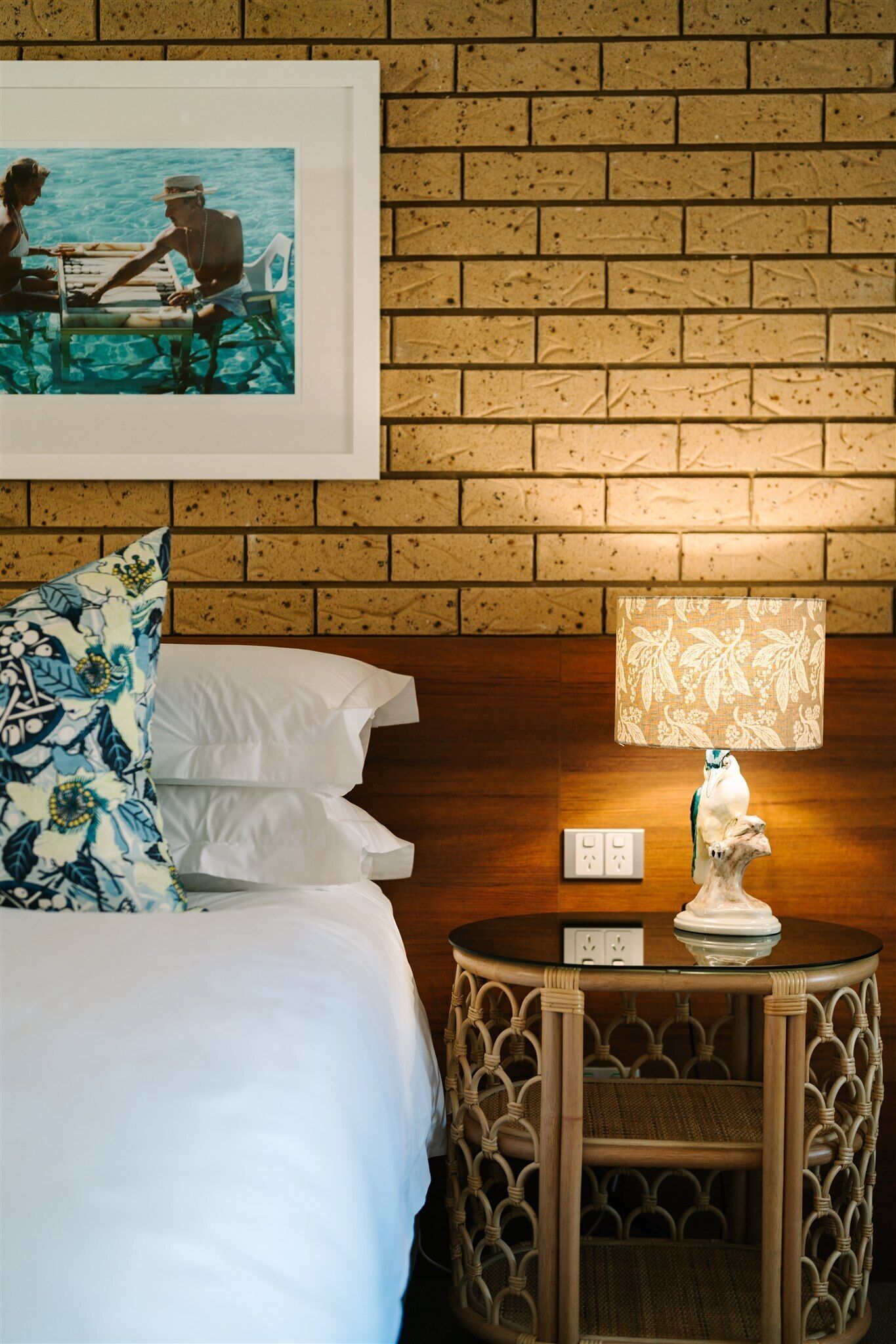 A hotel room with a brick wall, a framed photograph, a round table with a lamp, and a bed with white bedding.