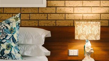 A hotel room with a brick wall, a framed photograph, a round table with a lamp, and a bed with white bedding.