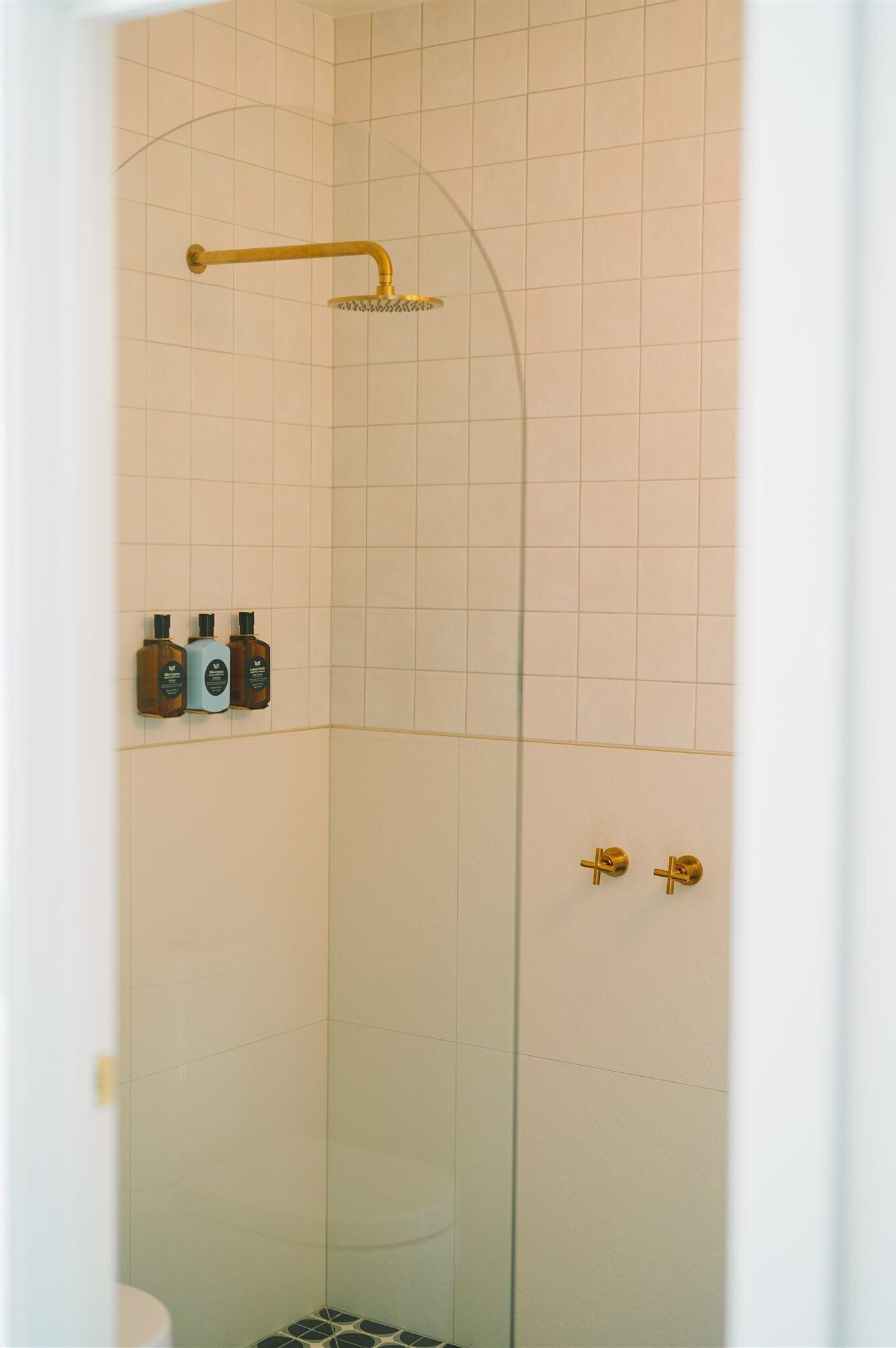 A glass-enclosed shower with a gold showerhead and control panel, a white tiled wall, and a small shelf with three bottles.