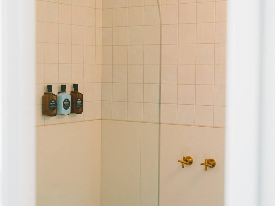 A glass-enclosed shower with a gold showerhead and control panel, a white tiled wall, and a small shelf with three bottles.