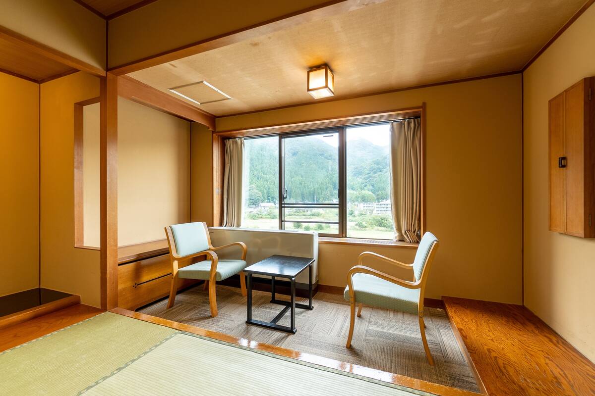 Una habitación con suelo de madera, un futón de tatami, una pequeña mesa, dos sillas y un ventanal con cortinas.