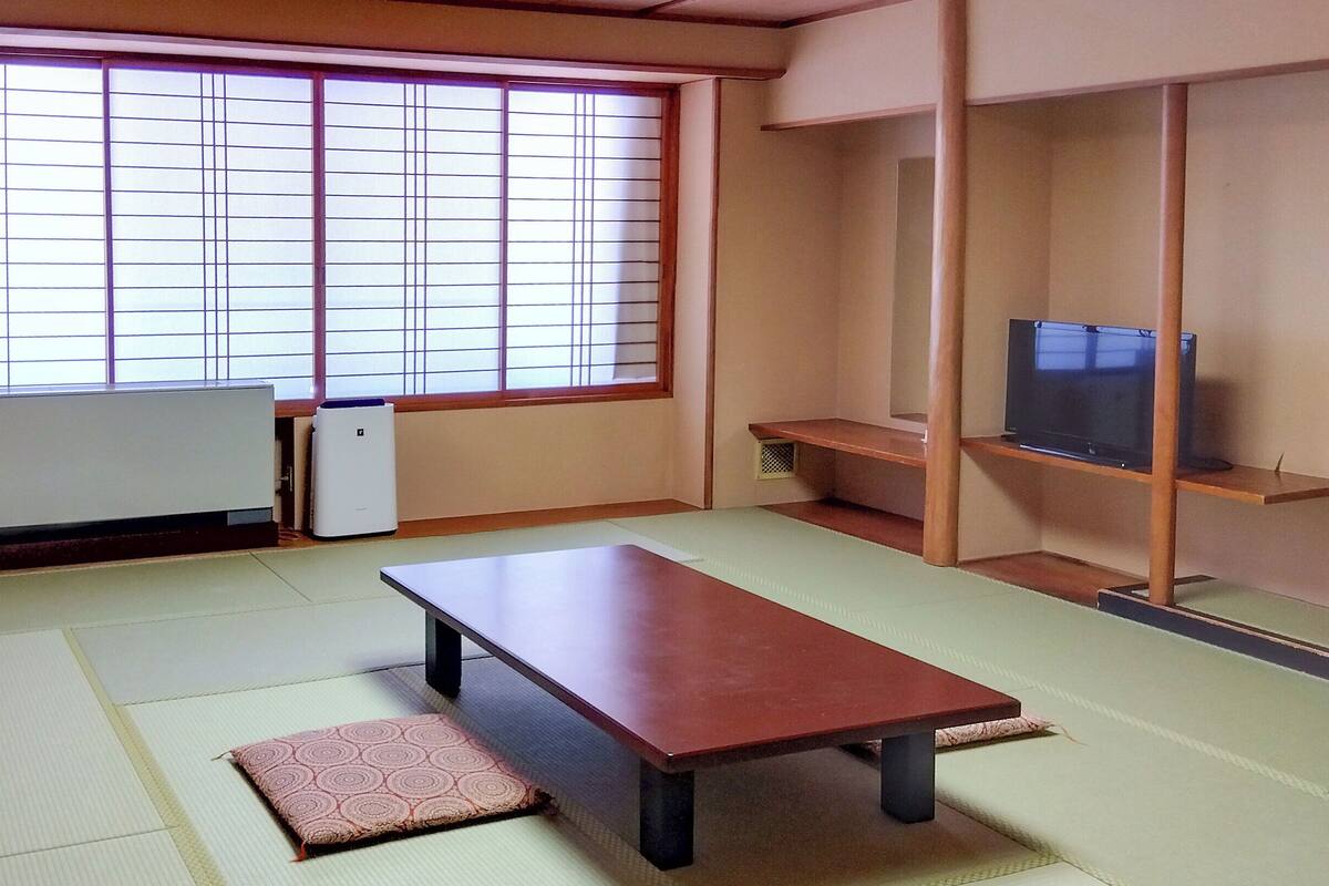 Una habitación japonesa tradicional con suelo de tatami, una mesa baja, un televisor en un estante de madera y una puerta corredera con paneles shoji.