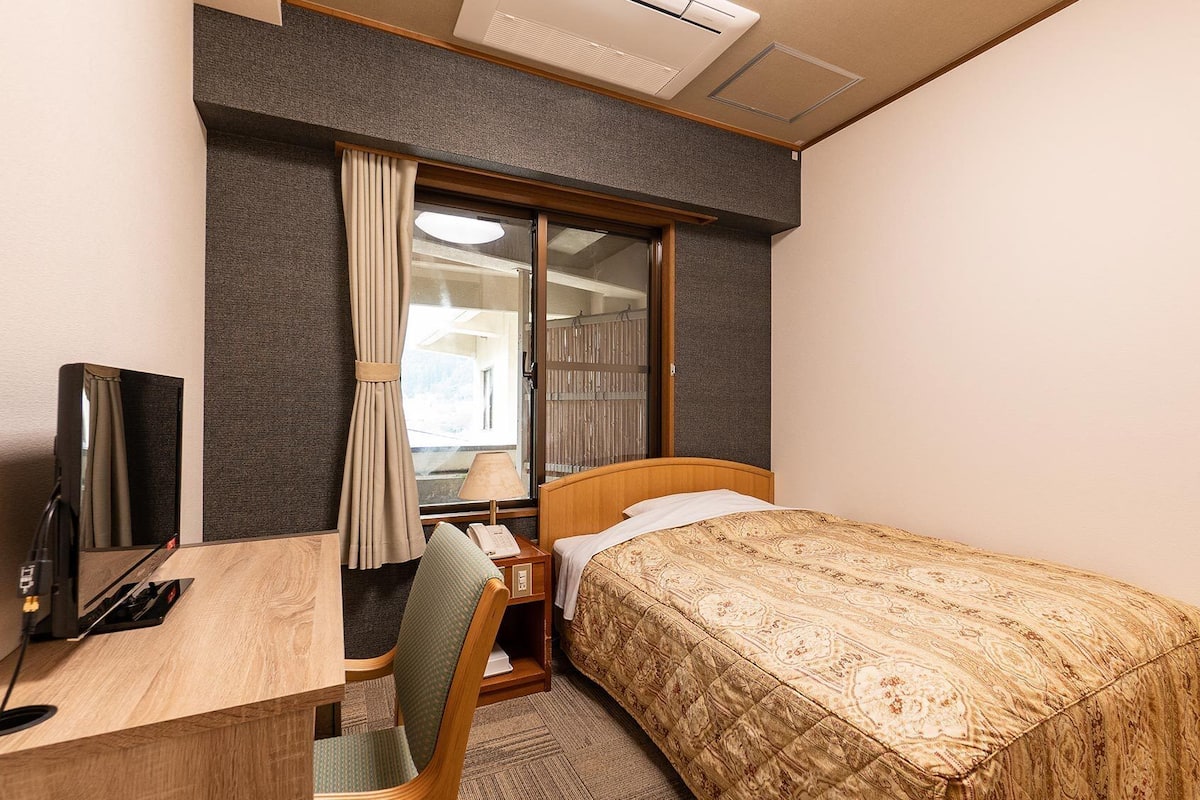 Habitación de hotel con cama, escritorio, silla, televisor y ventana con cortinas.
