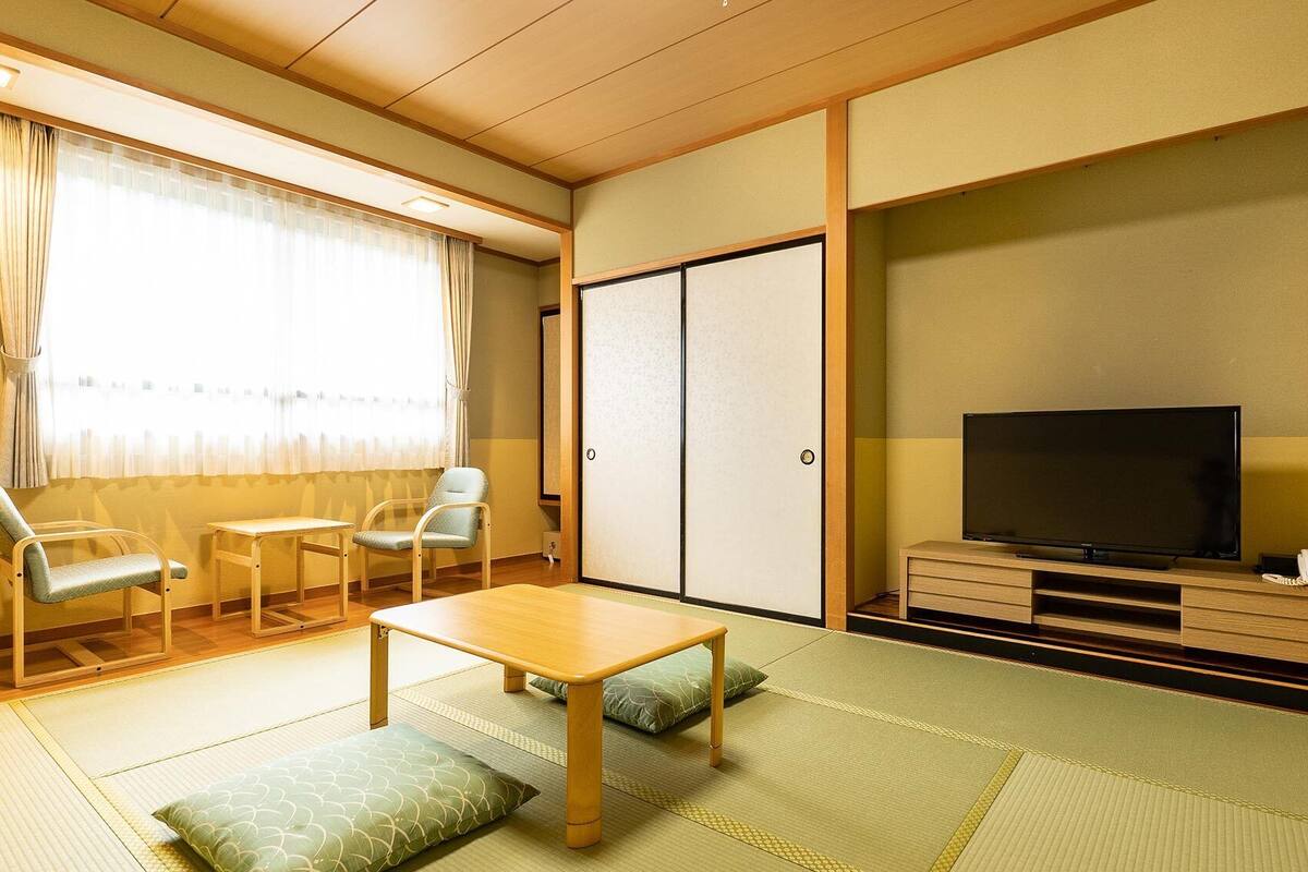Una habitación tradicional japonesa con suelo de tatami, una mesa baja, dos sillas, un televisor y una puerta corredera.