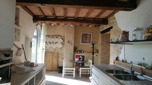 A rustic kitchen with a stone wall, wooden beams, a dining table with chairs, and a built-in sink.