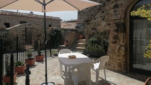 An outdoor patio with a stone wall, a white table, white chairs, a large beige umbrella, and potted plants.