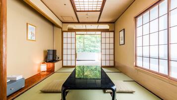 Et traditionelt japansk rum med tatamimåtter, et lavt bord, et fjernsyn på et træstel og udsigt til grønne områder gennem en shoji-skærm.