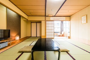 Ein traditioneller japanischer Raum mit Tatami-Bodenbelag, einem schwarzen Tisch, einem Fernseher und einer verschiebbaren Tür zu einem weiteren Raum.