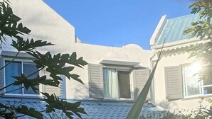Un bâtiment doté d’un toit recouvert de tuiles, d’un porche avec des balustrades blanches et d’un jardin où poussent des plantes aux grandes feuilles.