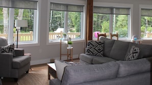 A spacious living room with a gray sofa, a coffee table, and a large window overlooking a deck. - 4-bedroom house in Stanhope - walk to the beach! (York)