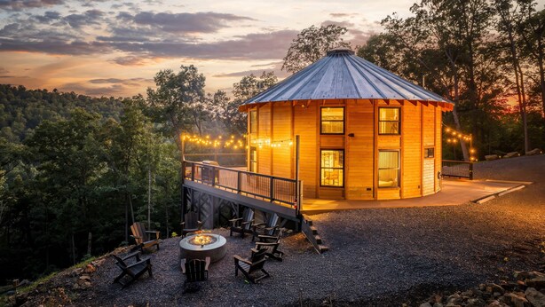 Una cabaña de madera con techo de metal, rodeada de árboles, con mobiliario exterior y una fogata.