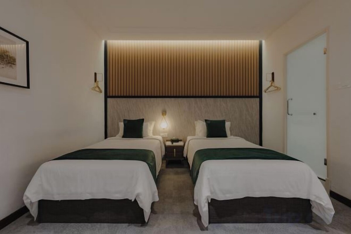 Quarto de hotel com duas camas, uma mesa de cabeceira com abajur, luminária na parede e uma porta.