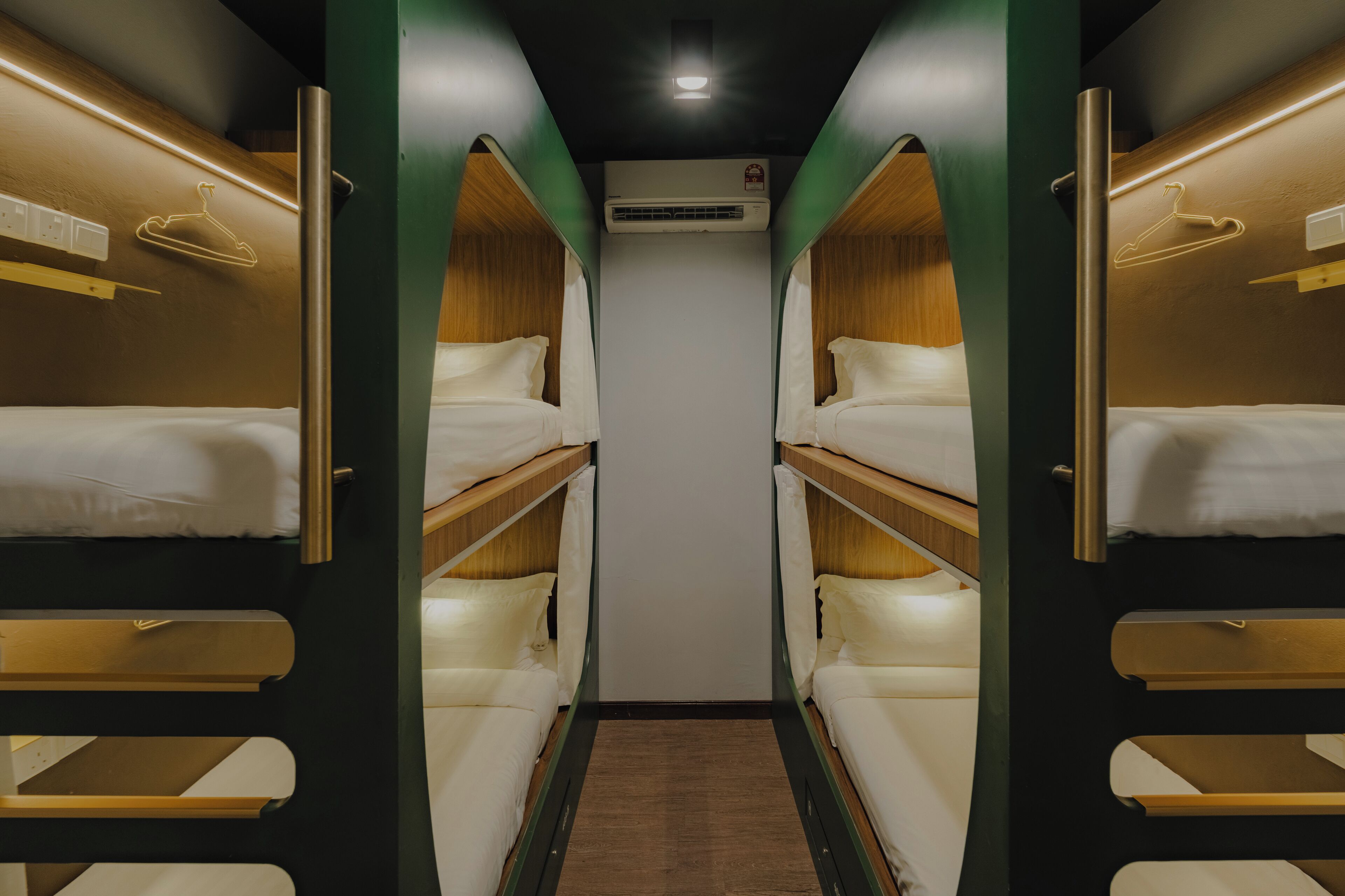 A narrow, well-lit corridor with bunk beds on either side, a central air conditioning unit, and a wooden floor.
