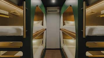 A narrow, well-lit corridor with bunk beds on either side, a central air conditioning unit, and a wooden floor.