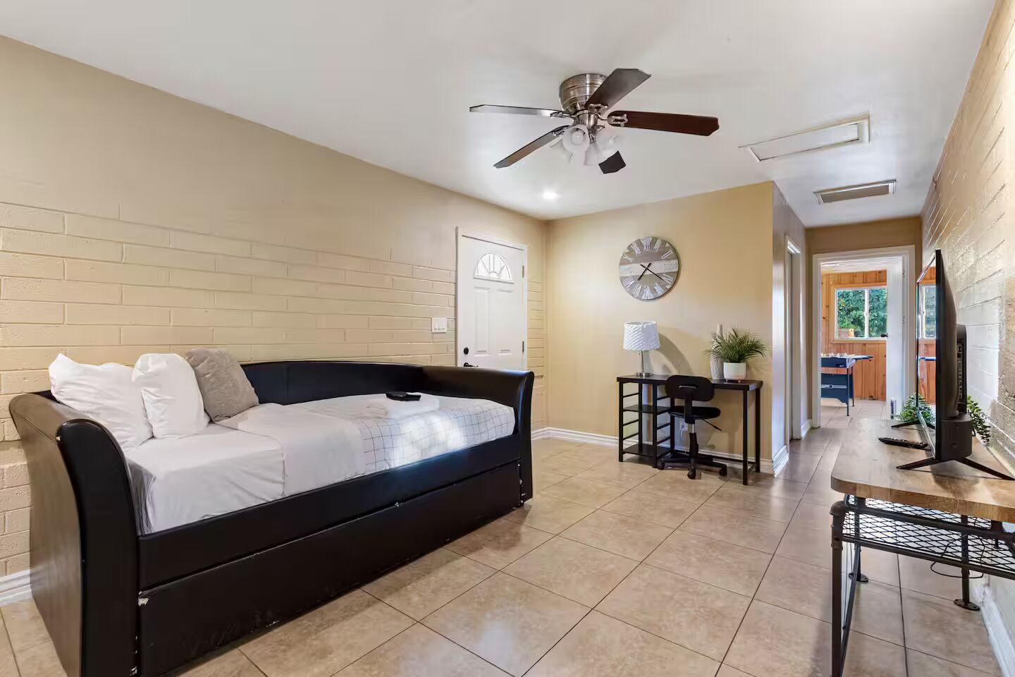 Une pièce moderne avec un canapé noir, un bureau avec une chaise, un ventilateur de plafond et une horloge au mur.