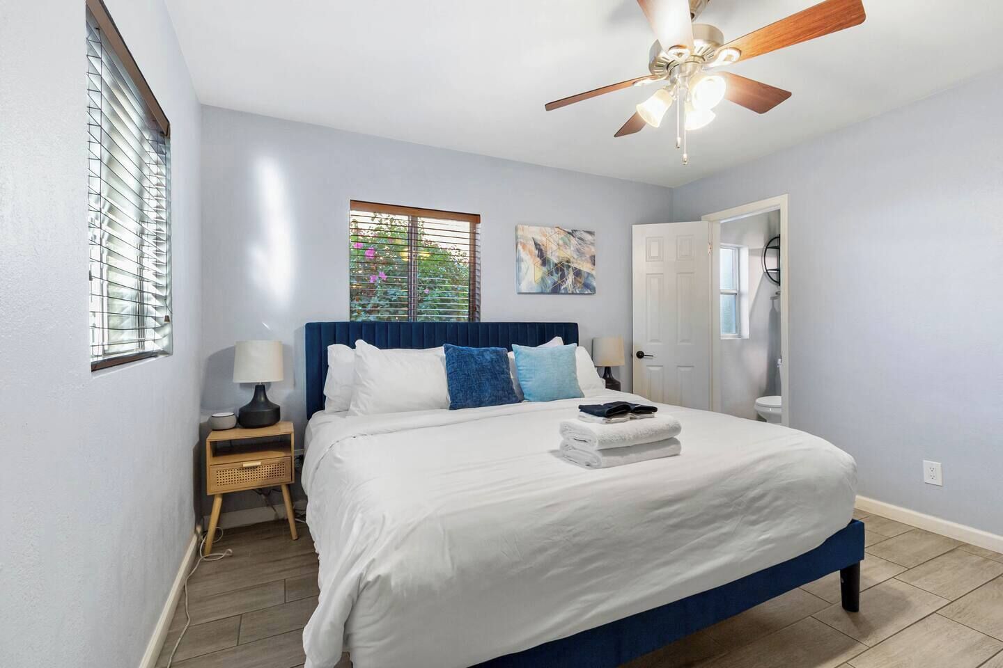Une chambre à coucher comprenant un lit, un ventilateur de plafond, une fenêtre avec des stores, une table de chevet avec une lampe et une porte donnant accès à une salle de bain.