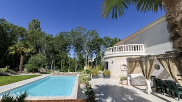 A pool area with a wooden deck, a white building with a balcony, and surrounding greenery.
